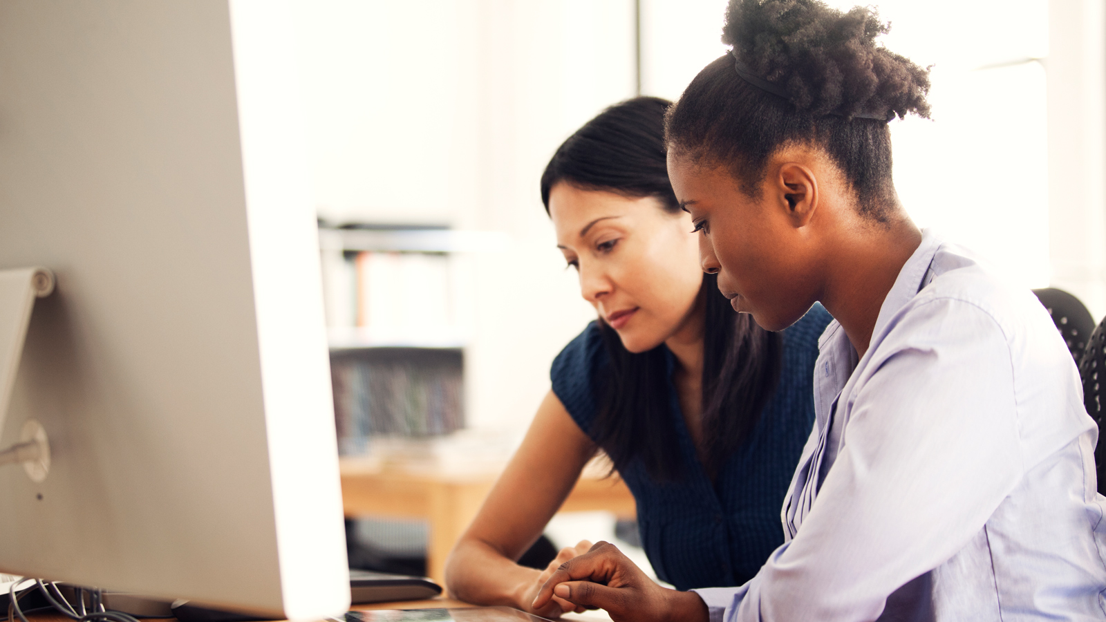 Two women sit in front of a computer and discuss content on screen.