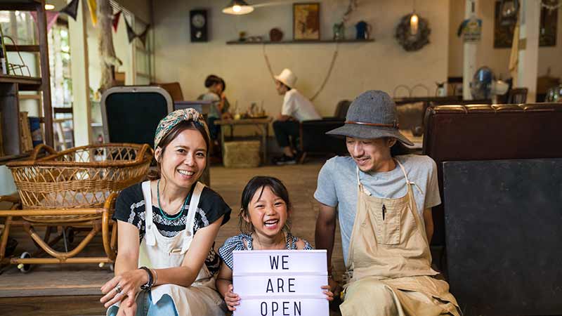 A husband wife and their kid sitting with a placard saying "We are open" in front of their shop.